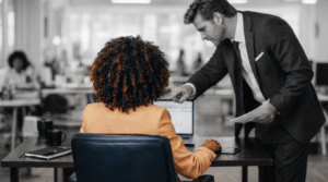 A manager leaning over an employee’s desk in a tense workplace interaction, highlighting pressure and power imbalance in a corporate environment.