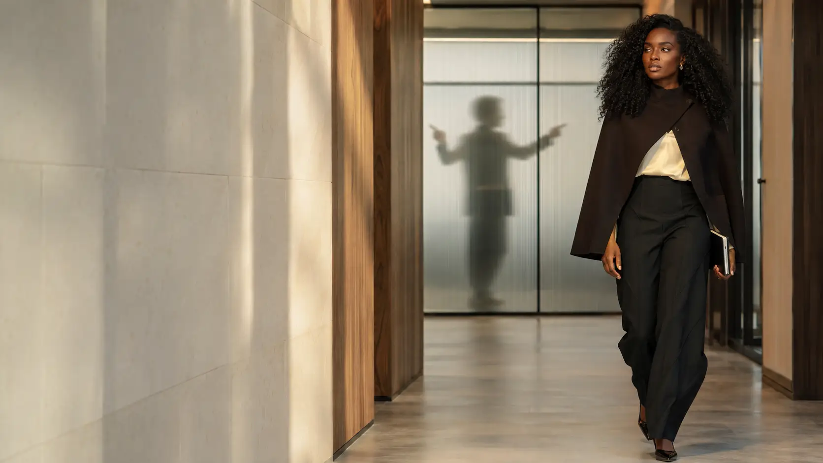 Black professional woman leaving a meeting room while a manager gestures behind frosted glass